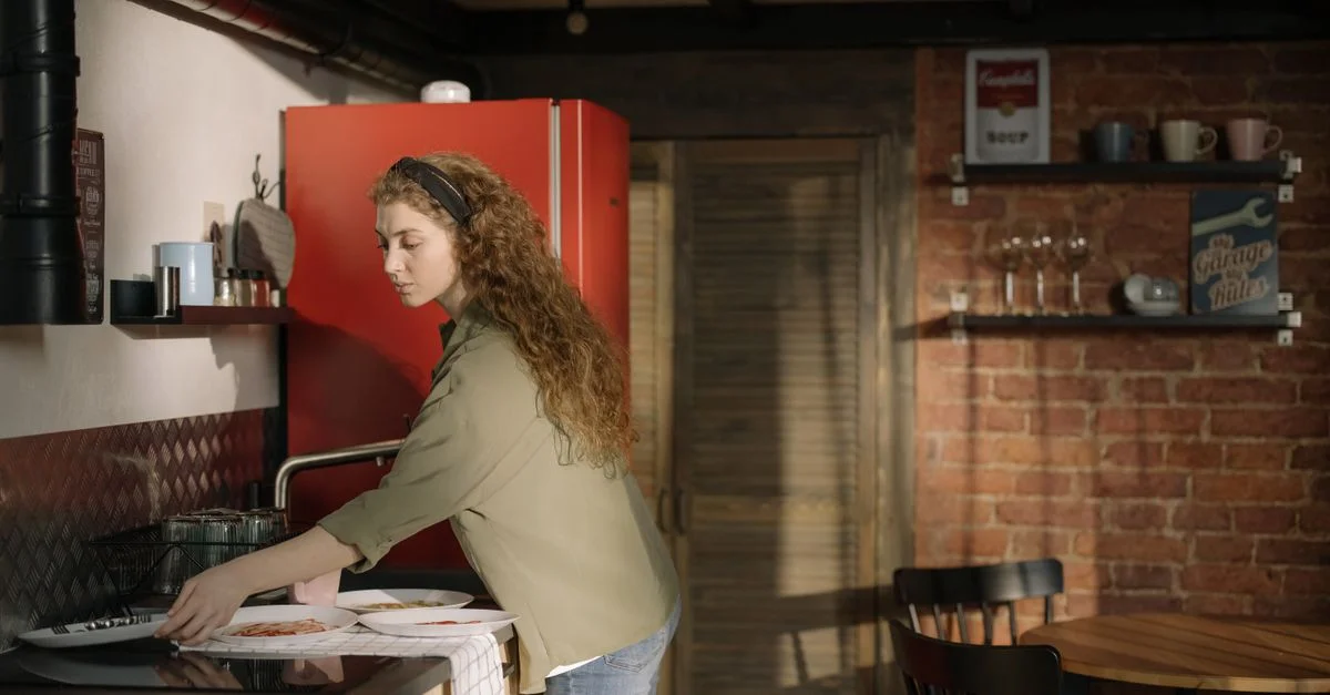 Une femme range de la vaisselle dans une cuisine chaleureuse avec un réfrigérateur rouge et un mur en briques.