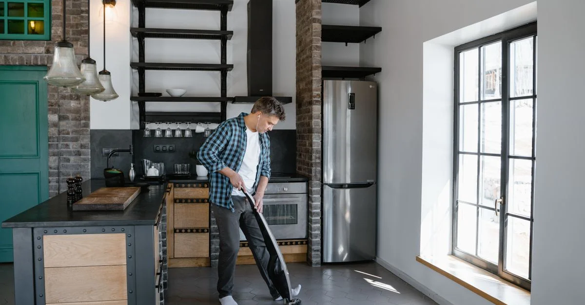 Un homme nettoyant la cuisine moderne avec un aspirateur, mettant en valeur le design intérieur contemporain.