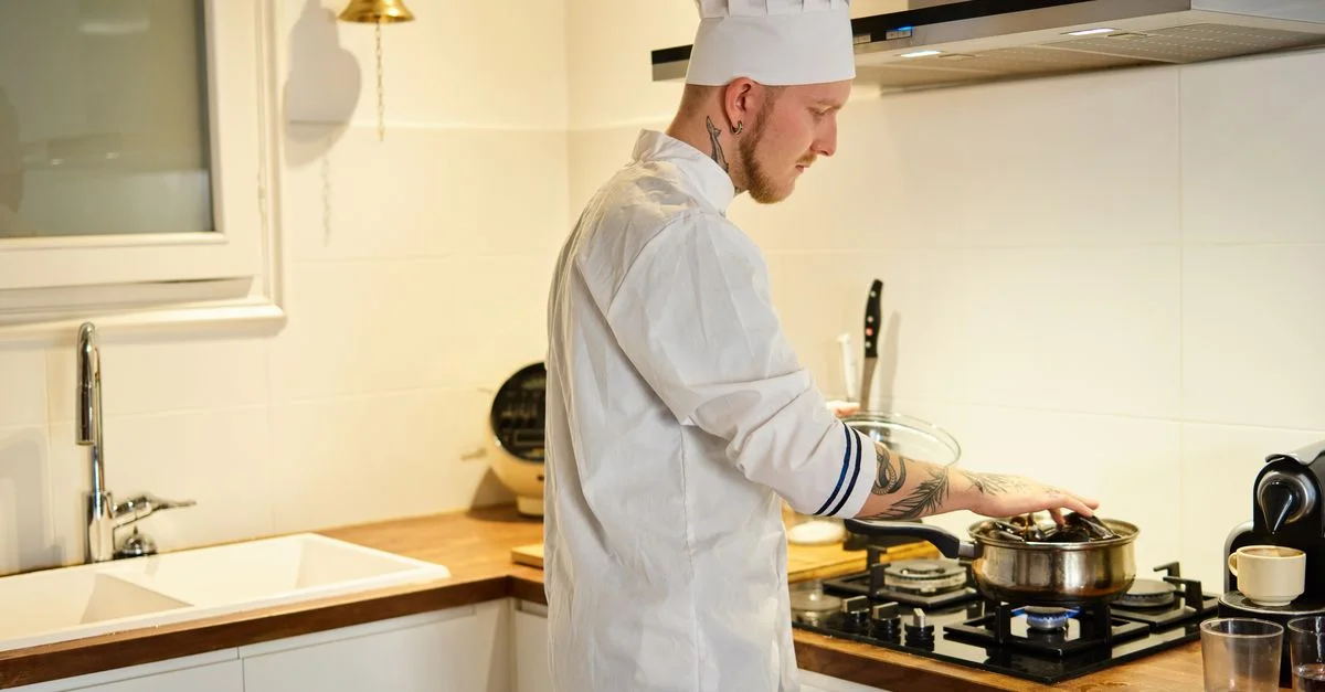 Un chef tatoué portant un chapeau cuisine dans une cuisine moderne, se concentrant sur une casserole sur le feu.