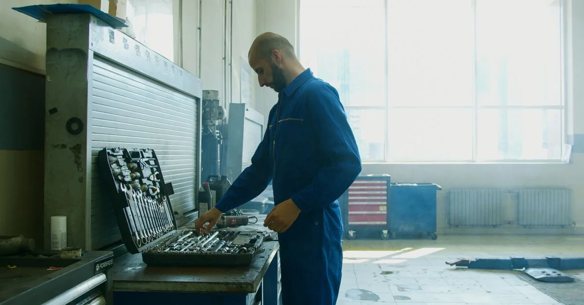 Un mécanicien inspectant des outils dans un atelier, portant une combinaison bleue avec une lumière naturelle vive provenant des fenêtres.