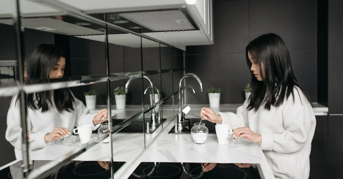 Une femme dans une cuisine moderne en train de mélanger du sucre dans une tasse avec des reflets miroitants.