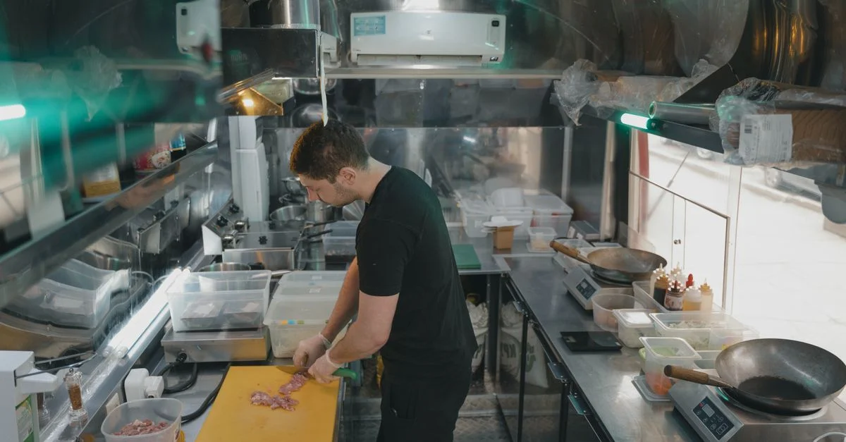 Un chef travaille dans une cuisine industrielle moderne, tranchant des ingrédients sur une planche à découper jaune.