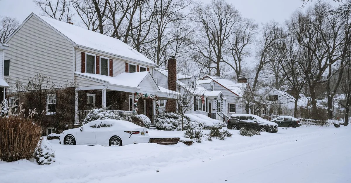 Une rue résidentielle tranquille recouverte de neige avec des maisons, des voitures et des arbres sans feuilles en hiver.