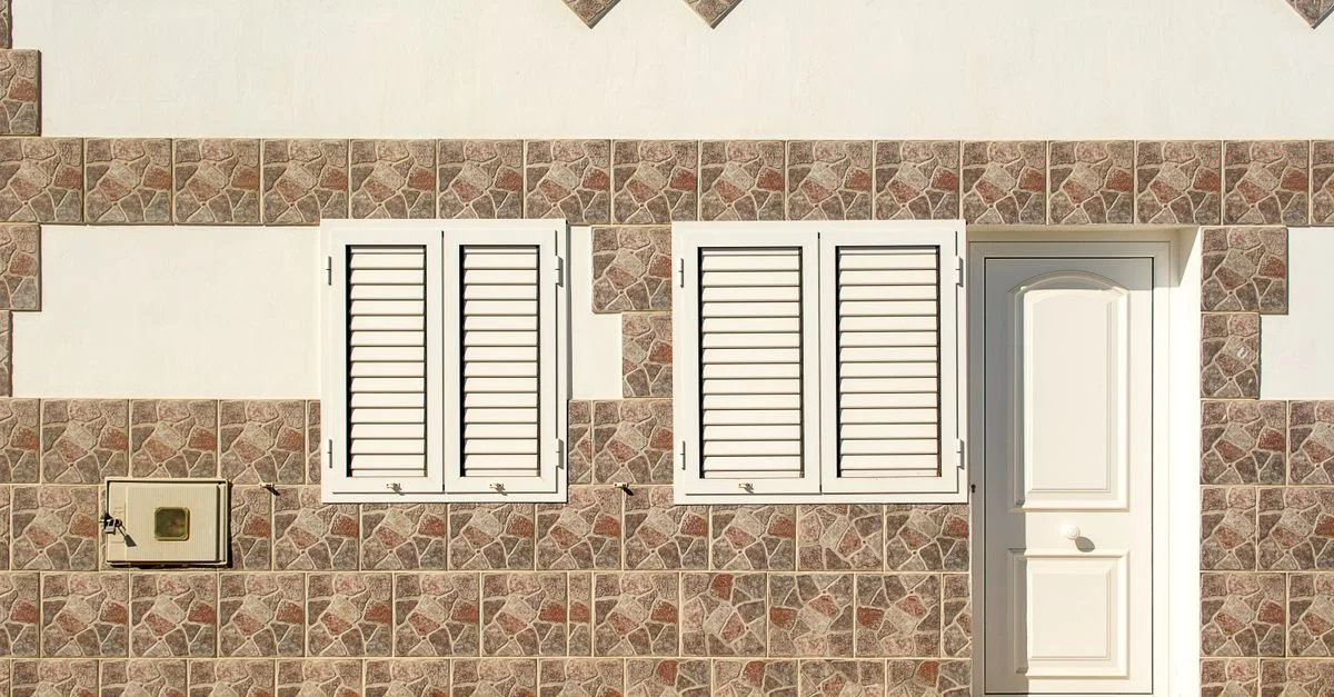 Gros plan sur une façade de bâtiment avec deux fenêtres à volets blancs et une porte blanche intégrées dans un mur en mosaïque de carreaux.