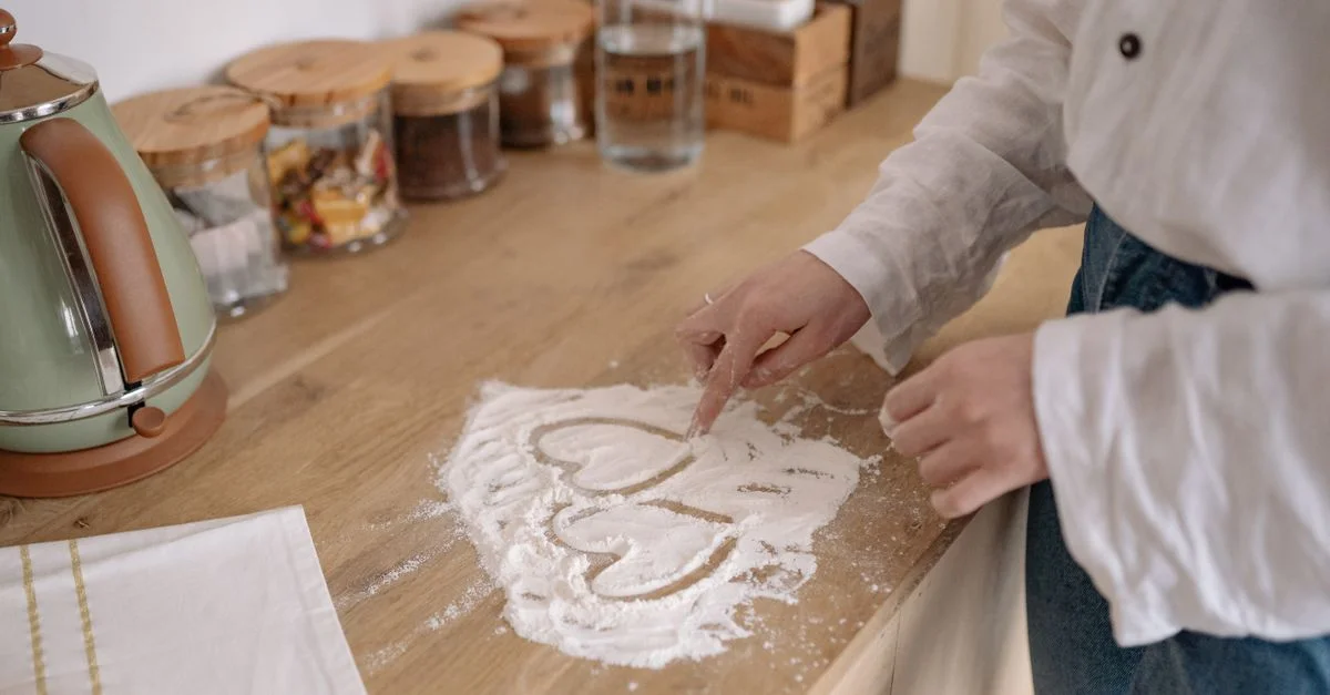 Une personne utilise son doigt pour dessiner un cœur dans la farine sur un comptoir de cuisine en bois.