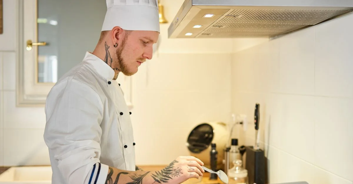 Un chef tatoué portant un uniforme et un chapeau blancs cuisinant dans une cuisine contemporaine.