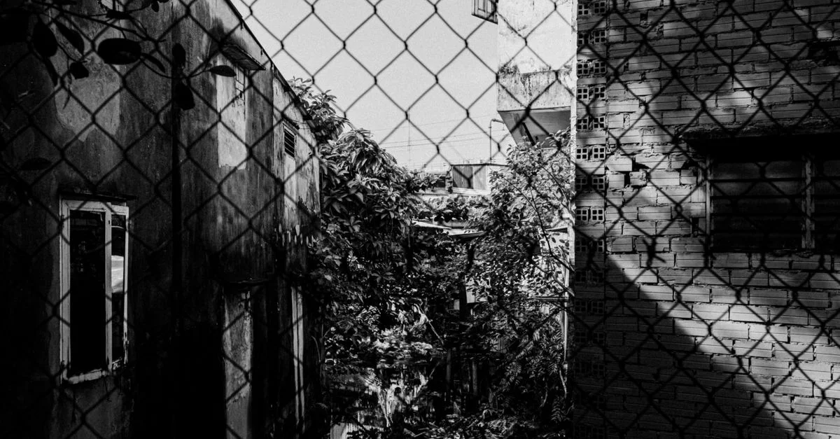 Photo saisissante en noir et blanc d'une ruelle urbaine, avec une clôture en grillage projetant des ombres.