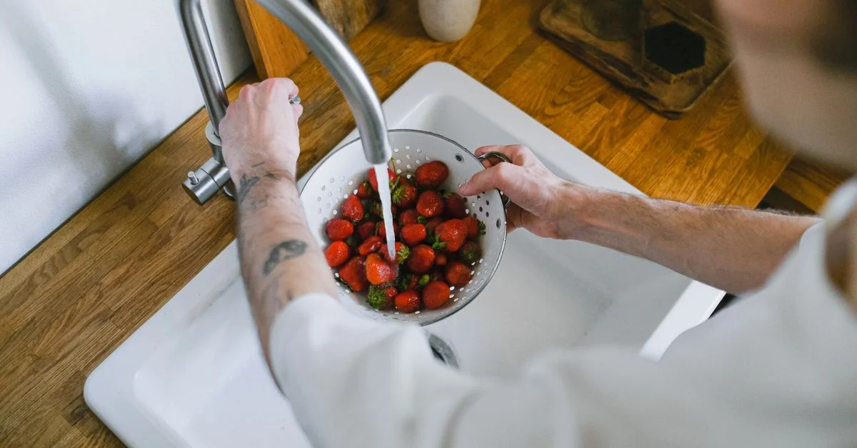 Une personne lave des fraises fraîches dans un évier de cuisine à l'aide d'une passoire. Un angle de vue en hauteur capture le processus.