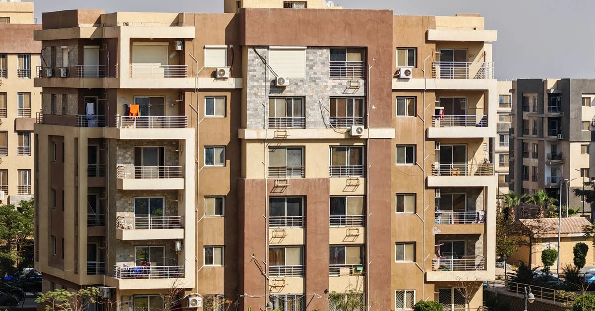 Scène urbaine ensoleillée montrant l'extérieur d'un immeuble d'appartements moderne avec balcons et verdure.