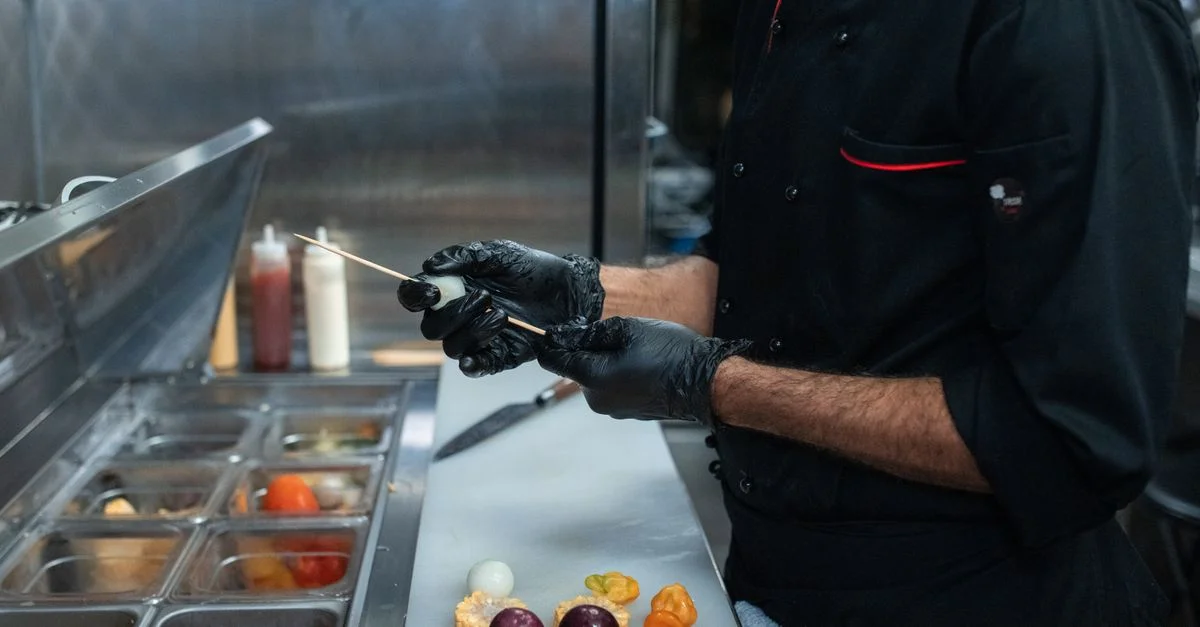 Un chef portant des gants noirs pique des légumes dans un cadre de cuisine commercial.